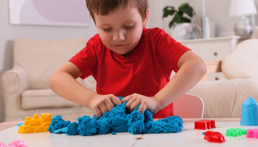 Cute little boy playing with bright kinetic sand at table in room