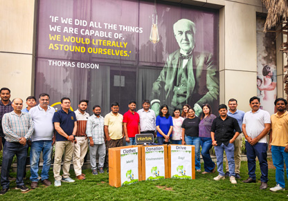 A large group of Intertek India colleagues stands behind marked clothes donation boxes. 