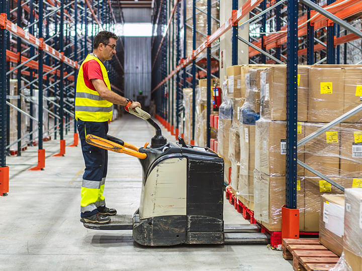 Worker wearing yellow safety vest in warehouse picking up a pallet of products with an electric pallet jack