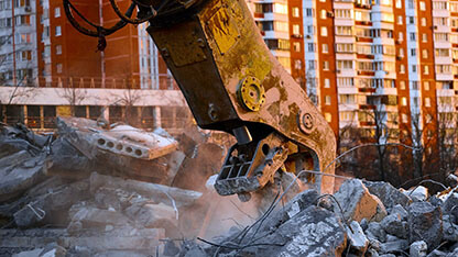 Concrete crusher on excavator rigging works with pile of reinforced cement leftovers for recycling at demolition site at sunset