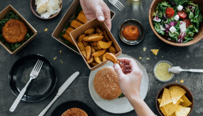Hands sharing fries over table with burger, salad, chips and assorted dishes during meal.