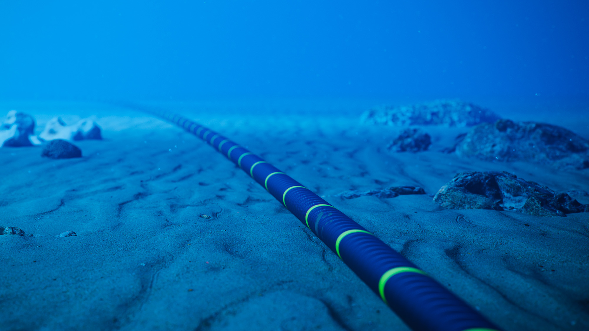 Underwater view of a subsea cable stretching across the sandy ocean floor, disappearing into the blue distance. Rocks and seabed features surround the cable, highlighting offshore infrastructure in a deep-water marine environment.